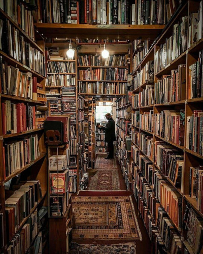 Photo of a used books store in Edinburgh packed from top to bottom with books. At the end of a bookshelf corridor, we see a man standing with an open book in his hands.