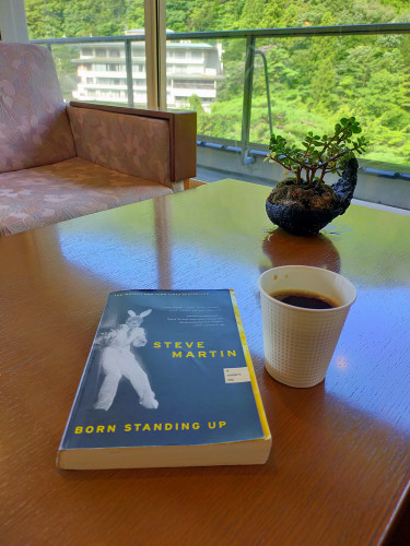 Photo is of a wooden table. On the table is a black paperback book with a white image of a young white Steve Martin in a white suit & pants & bunny ears. To the right is a white up if black coffee. There is a tiny green bonsai on the table in a black planter  In the distance is a pink cushioned love seat. Out the window is tons of green trees and a white hotel