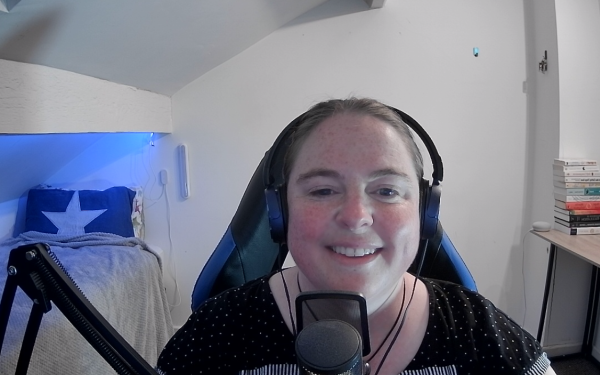 Becci is sitting in front of her computer. In the background you can see her office room with books and a made bed. She is smiling and wearing headphones, and there is a chunky microphone in front of her.