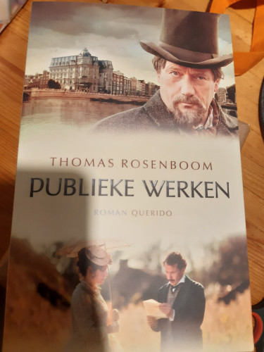 Book cover, with two images from the film. Above the title and author: the site of the hotel, with the main character's house. In the foreground a man with a top hat and a goatee looks into the camera. Below: a man and woman. She holding a parasol, he reading a letter 