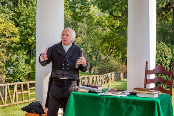Actor portraying James Madison stands in front of a table scattered with books and writing instruments. A tall chair sits to the right. A tri-cornered hat sits on a leather bag in front of the actor. The setting is outside under a temple with large white columns. 