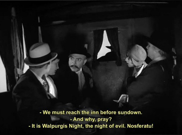 A scene from a black and white film. It shows five or six travelers in the back of a horse-drawn carriage, having an intense conversation. Someone says, "We must reach the inn before sundown." Someone else replies, "And why, pray?" The first man replies, "It is Walpurgis Night, the night of evil. Nosferatu!"