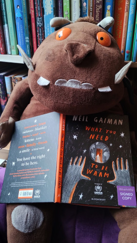 Cuddly Gruffalo plushy holding a copy of What You Need to be Warm by Neil Gaiman, on an armchair in the Edinburgh Bookshop
