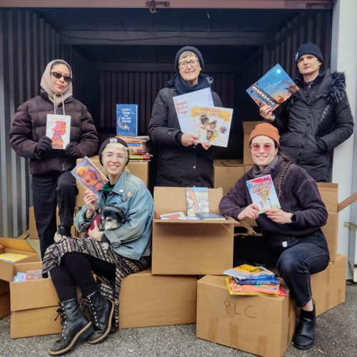 Five humans and one canine sit and stand among an array of cardboard boxes. Some of the boxes are open revealing the contents inside: picture books and middle grade novels. Each person is dressed warmly and holds at least one book with the cover facing the camera.