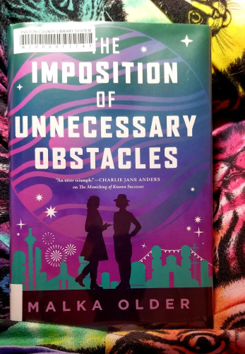 A book titled The Imposition of Unnecessary Obstacles, Malka Older's sequel to The Mimmicking of Known Successes, rests on my lap. I'm wearing pajamas with imprints of cats set against psychedelic colors. 