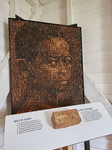 A mosaic portrait of a young African boy made from pieces of brick. Beneath the mosaic is an exhibit panel that has a 2 short paragraphs about enslaved people making bricks by hand. A handmade brick is affixed to the panel, featuring fingerprints. 