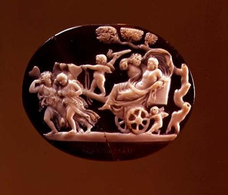 Cameo of Dionysos lounging in his chariot, his cloak billowing over his head. The chariot is small, with only one seat. A satyr is walking beside the chariot, embracing Dionysos and looking lovingly at the reclining god. Eros is standing on the holding up the torch with which he lights the hearts of gods and men with desire. In his other hand he is holding the reins to the chariot that is drawn by a tam of maenads or maybe Psyches, since they seem to be winged too.
The scene is carved in sardonyx against a dark, onyx background.