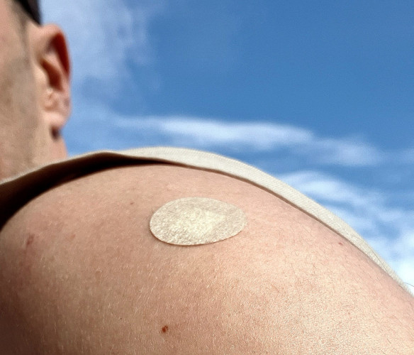 Close up of my shoulder against a background of clouds. On my shoulder is a little patch on the spot where I was vaccinated.