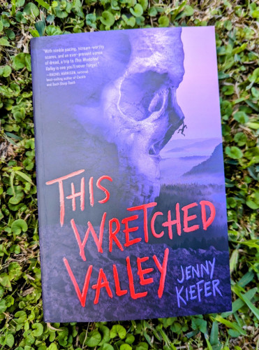 Paperback of THIS WRETCHED VALLEY by Jenny Keifer. The cover is purple with a rock outcropping shaped like a human skull in profile with a single person dangling from the tip of its "nose" and the vast wilderness below them.