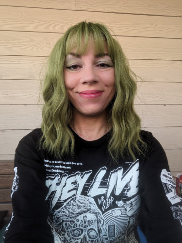 Selfie of a middle aged woman with dark eyes and green eyeshadow wearing a green wig and a They Live shirt