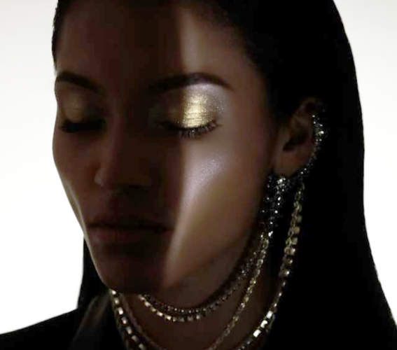 An artistic and alluring tight shot of a Black woman’s face in partial shadow. Sparkling beads hang underneath her chin and are attached to both ears. Her eyes are closed and her eyelids are dusted in gold eyeshadow.