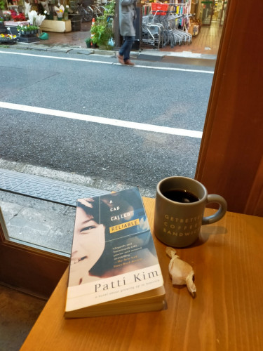 The photo is of a small wooden table against a window. the thin paperback book has a photo of Korean American girl's half face smiling. To the right is a grey coffee mug of black coffee with Get Better Coffee Sandwich on it. Below the mug is a tiny cookie wrapped in white paper. Outside you can see a street and a Japanese man walking by but his face is not visible