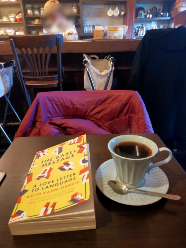 The photo is inside a cafe. The yellow paperback book with various block letters from various different alphabets spread around. To the left is a white porcelain mug of black coffee on a similar currogated saucer. A metal silver spoon is in front. On the other side of the table is a purple jacket. In the distance is the counter behind which is the coffee master whose face is blurred but is wearing a beret made from an old tan raw coffee bean bag