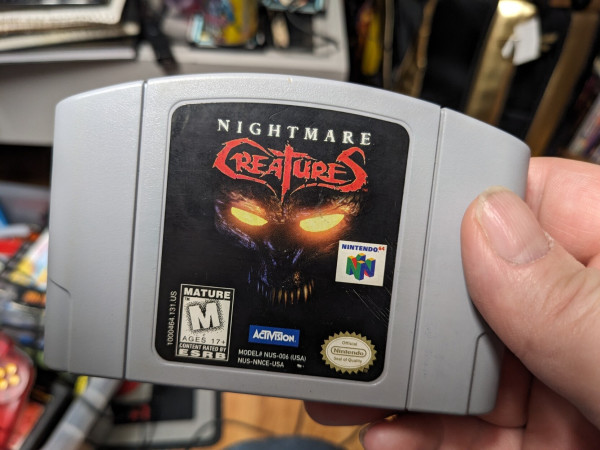 A photo of the Nightmare Creatures N64 cartridge. It has a black label with the game's logo above a shadowy spiky-toothed skull-like face with glowing orange eyes