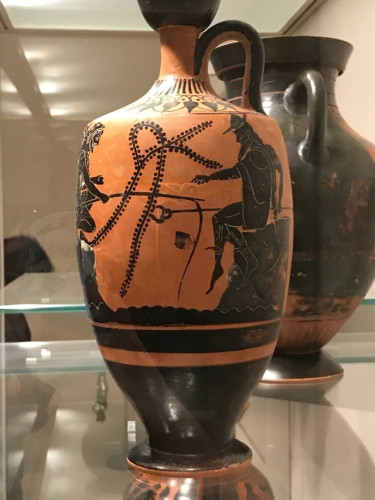 Detail of a terracotta lekythos (a type of vase) with a black-figure vase painting of Herakles and Hermes fishing, each perched on a rock. Herakles tries his luck with a rod while Hermes just sits there, gesticulating to the right.