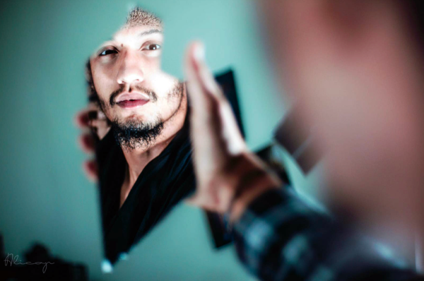 A guy stares at his reflection in a broken piece of mirror he holds in his hand. The twist is that the image is shot from over the guy’s shoulder, giving us his perspective.