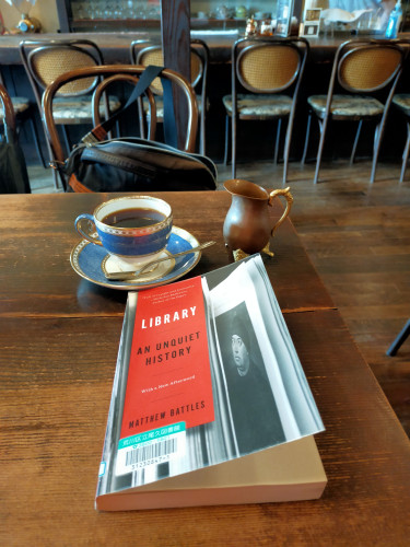 Photo is of the inside of the coffee shop. In the distance are the backs of 4 rattaned chair backs against a counter. In the foreground is the paperback library book which has a red vertical rectangle of the title & author on the left side and a black and white image of book pages of a white man looking up. Behind the book is a white and blue saucer and cup of black coffee with a silver spoon facing left perpendicular on the saucer. To the left is a brown miniature milk pitcher. A brown shoulder bag sits in the wooden rattan chair on the other side of the brown wooden table