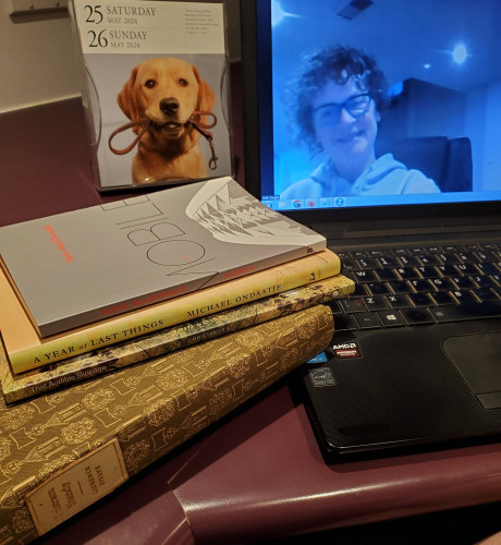Me, on screen, getting ready for a silent book club zoom meeting - a dog calendar showing the date (Saturday, May 25th) and a stack of books by Tanis MacDonald, Michael Ondaatje, Margaret Christakos and Laurence Sterne sit next to the computer