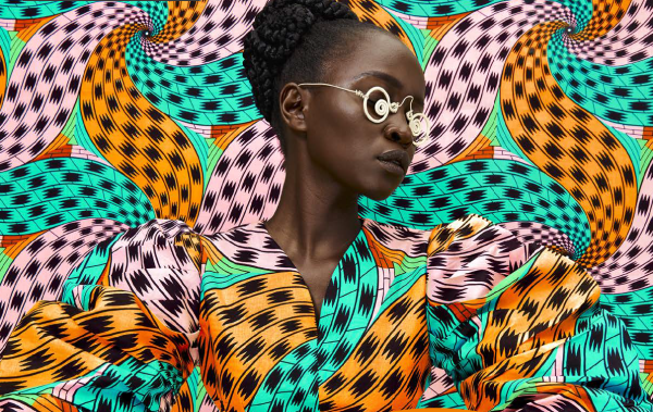 A woman in an African print dress is posing in front of a colourful background. The background and dress have an identical pinwheel type pattern in light pink, turquoise, and orange with black accents. The woman is wearing wire glasses.