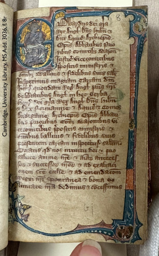 Photo of a leaf in a medieval manuscript: folio 8 recto in Cambridge, University Library, manuscript Additional 3036. At the bottom of the image, the tip of a woman’s finger holds the page flat and reveals the book to be downright minuscule—smaller than a pack of cards! The displayed page bears 24 lines of Latin text copied in dark brown ink, with the first 7 lines indented to accommodate an enlarged decorative initial ‘E’ (for ‘Edwardus’). A painted border in blue, pink, and gold extends from the initial, encircling the text on 3 sides. The initial itself, in blue on a pink ground, is infilled with gold and features a depiction of a king, crowned and enthroned. The tiny monarch is clean-shaven and sits turning slightly towards the right, as if reading the start of the text. He grasps a large sword with one hand, holding it upright. 