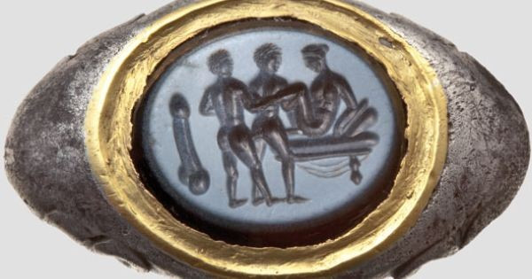 A Roman silver ring with erotic intaglio depicting two men and a woman engaging in a "love chain" with the woman reclining on a couch, receiving one of the men and the other man fucking him in the butt. A large phallus is depicted on the left to the scene, possibly for good luck.