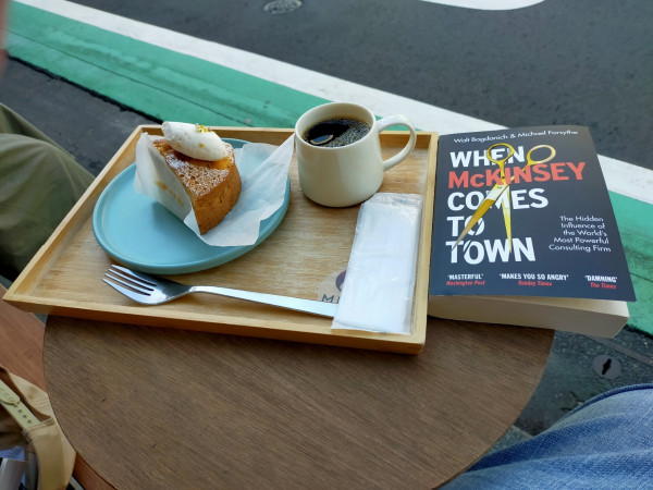 Photo is outside of the coffeeshop. On a small round wooden table is the black paperback book with golden scissors & McKINSEY in red & the rest of the words in white. To the left is a wooden tray on which is a metal fork, a white napkin in plastic, a white mug of black coffee, a turquoise tray with a slice of brown sugar frosted cake & an oval dollop of cream on top, the whole thing on white translucent paper. Beneath the table on the street is a green stripe outlined by white line