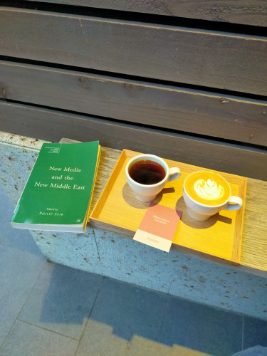 The image is a photo of a narrow wooden bench on a concrete block. The bench is against a dark wooden fence. The book is green with the title listed in the toot. It is on the left edge. To the right is a wooden tray with a white mug of black coffee and a white mug of a flat white with tan foam & white rosette. Between them is a mostly pink card with white words Myanmar Genius and a smaller white section with pink ALLPRESS.