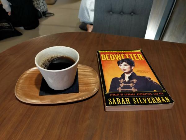 Photo is of a cafe table on which is the paperback book with a headshot of white comedian Sarah Silverman with her black hair tied up in a bun wearing a regal European military style black jacket with gold rope-ish designs horizontal in front. To the left is a white tea cup mug of black coffee on a small oval balsa wood -y tray