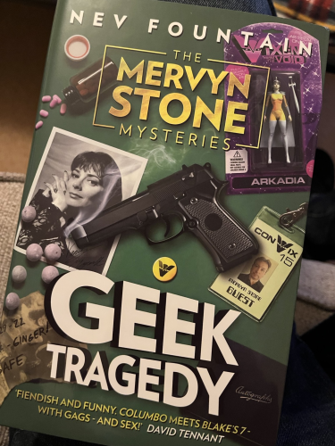 Front cover of Geek Tragedy. The cover shows a gun alongside paraphernalia from a Sci-fi convention including a toy figurine, a guest pass and a black and white photo of an actress.