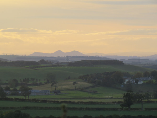 A muted, sunset landscape. A faintly pinkish sky above a blue and purple horizon, layers of hills fading into the distance. Three distinct peaks of the Eildon Hills stand alone, higher than the rest. In the nearer landscape, the valley is green and shadowed, lined with dark hedges and sprinkled with clusters of trees. An old, stone farm sits in the bottom of the valley, with long, low barns and a solitary Scots pine towering over it. To the right, on the side of a rise and partly hidden by trees, nestles a small village of slate-roofed houses.