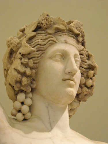 Detail of a marble sculpture of the god Dionysos. Close-up of his head photographed from the bottom left. He is crowned with a wreath of vine and ivy with small clusters of berries around the top of his head and big, heavy clusters of grapes hanging down by each side of his face. A serene smile suggests the benevolence of the god.