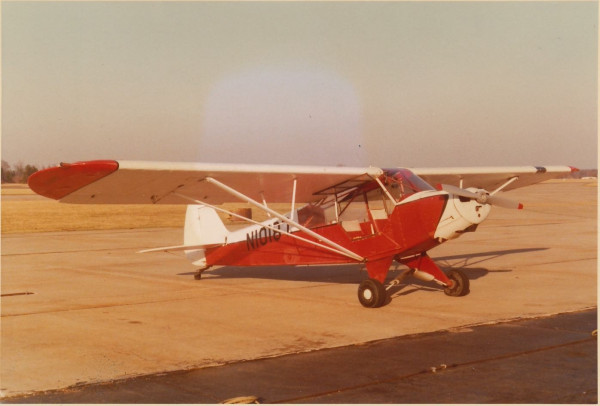 A red and white airplane, a Piper "Super" Cub (taildragger) sitting on an airport tarmac with the door open, no one inside.