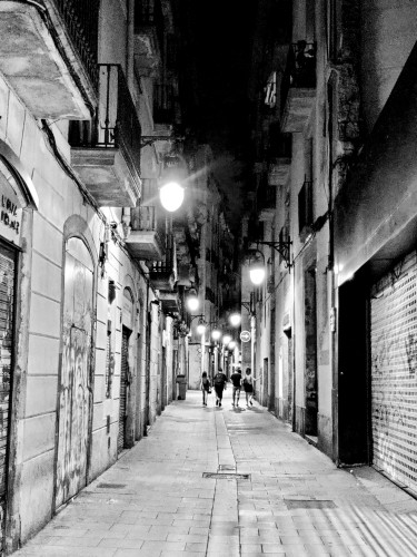 One of the narrow semi pedestrian streets in Barcelona, lit by lamps hanging from the buildings, alternating sides. Four people walk away from the camera in the distance. Graffiti is visible on the steel doors that have been rolled down to protect the closed storefronts. (Black and white.)