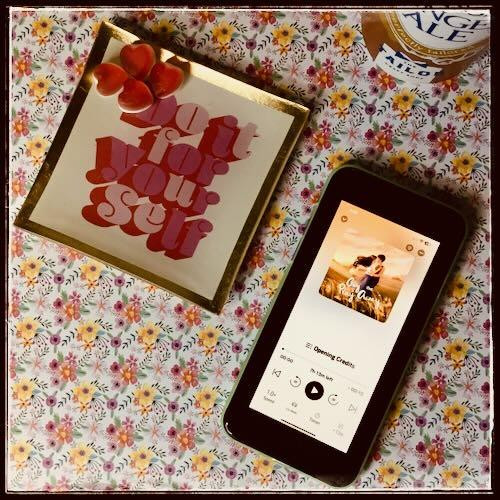 An iPhone with the cover of Gay the Pray Away, written and narrated by Natalie Naudus, on a flowery background. Next to the phone, in a small square plate with the words "do it for yourself" are several heart-shaped pieces of candy. On the other side, a glimpse of a can of ginger ale.