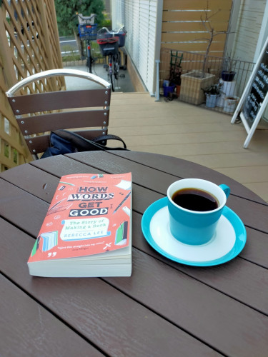The photo is the outside seating of a cafe. On a round brown faux wooden table is the red paperback book with a white Turquoise rimmed coffee cup & saucer of black coffee. Parked bicycles can be seen in the distance