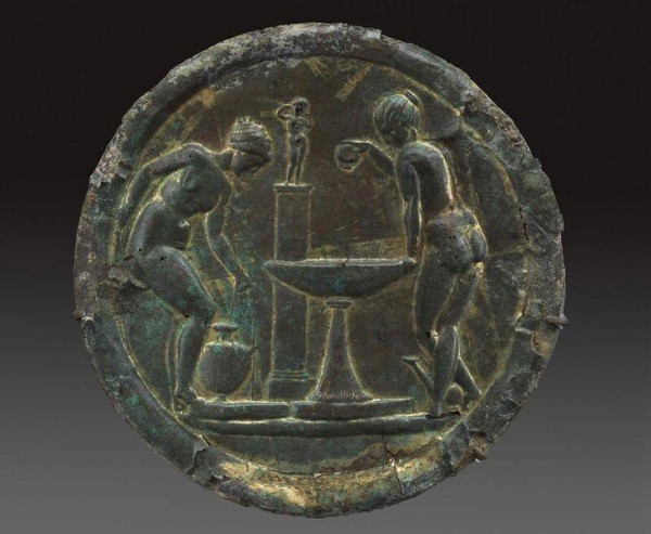 In the center is a large basin on a tapering pedestal. To the left, a nude woman with her hair wrapped in a cloth bends to pick up a water jar (kalpis) at the left. There are low bases or platforms below the kalpis and the basin. At the right, a nude woman with her hair pulled back tightly into a bun on the crown of her head leans on the basin with her left hand and pours oil into the basin from a bottle in her right. Behind her feet is an askos. Behind the basin is a pillar with mouldings above and below. The pillar is topped by a statue of a nude Aphrodite arranging her hair. There is a low base beneath the figures and containers.