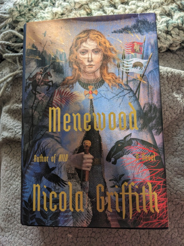A hardcover copy of Menewood by Nicola Griffith. A tall young woman with reddish hair, a Celtic-looking brooch, and a sword stands in the foreground.  In the background a battle rages with banners and men on horses.