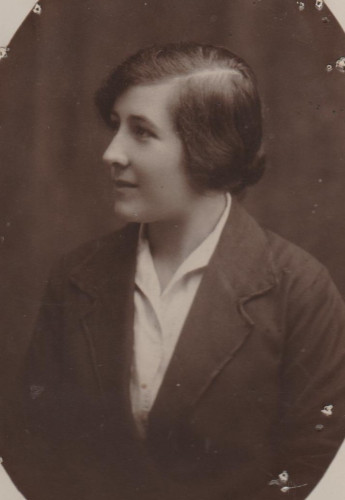 Dolors Martí Domènech in sepia toned black and white photo looing image left in profile. Dark dair in low bun, parting on her left. white blouse under dark jacket with lapels. young white woman smiing slightly.