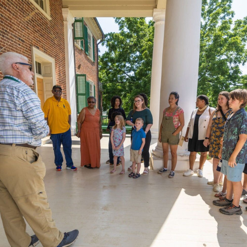 A diverse group of people stand in a semi-circle with their attention focused on the single man leading the group. Everyone is standing on the portico, or front porch, of the Madisons' historic home, known as Montpelier. It is a bright, sunny day. 