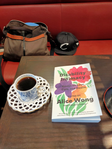 Photo is of a cafe table. The white paperback book with a flower illustration of red (top), purple (left middle), orange (bottom) & green leaves. To the left is a white mug (with blue unidentifiable illustration) of black coffee on a plate of woven ceramic. The table is brown and square. On the red bench opposite is a black baseball cap with a stylish C for the Cleveland Cavaliers. To the left is a brown shoulder bag with an orange stripe in the middle
