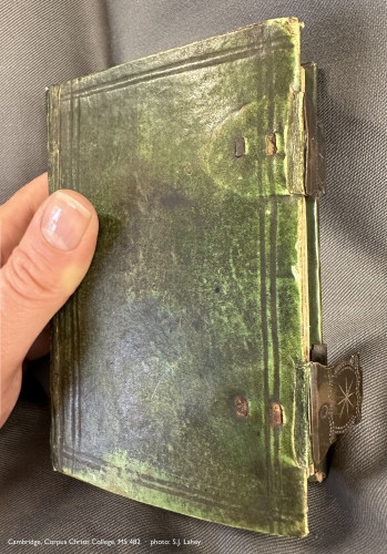 A very small book being held up over a grey fabric cushion. The book—Cambridge, Corpus Christi College, manuscript 482—is clad in a slightly scuffed green binding, with two engraved metal clasps visible at its fore-edge. At left, part of a woman’s hand is visible, grasping the book around its spine; her fingers are hidden behind the book, but her thumb rests atop its front board. The juxtaposition reveals the book’s compact size: its height is perhaps 7 times the width of the woman’s thumbnail. 