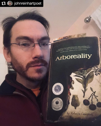 Poet John Reinhart holding a copy of Rebecca Campbell's Arboreality showing the Ursula K Le Guin Prize and Philip K Dick Award mediallions.