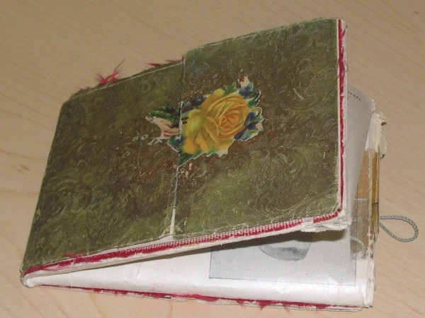 small folding album: patterned olive green with yellow rose

red stitched border

album cover is opened at an acute angle, offering a glimpse of interior

small blue-gray twisted fabric loop at right for securing cover