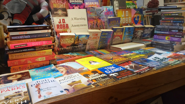 A display table full of modern paperback and hardcover books. There are books flat on the table, an upright rack with books facing forward, and a stack of books towards either end. No two books are the same but a few authors are represented by multiple titles. Too many books to list them all, a few of the visible names/titles are as follows:
Britney Spears/The Woman in Me,
J. D. Robb (x3), Anonymous/A Warning, John Ringo (x2), J. A. Jance, David Baldacci (x4), Vince Flynn, Janet Evanovich (x2), Tom Clancy (x2), John Sandford, James Patterson, John Grisham, John Bolton/The Room Where it Happened, Laura Graham (x2), Nicholas Sparks, Jodi Picoult (x2), Jack McDevitt, western, memoir, mystery, romance, young adult, horror, self-help advice, and more.