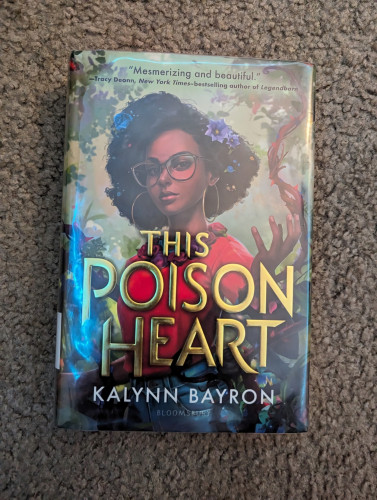 A book with a girl in a forest on the cover. She has vines wrapped around her and flowers in her hair. The book is titled in golden letters, "This Poison Heart." The author is Kalynn Bayron.