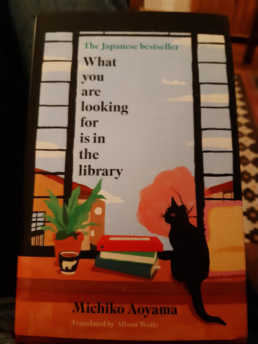 Book cover: an open window or door, sky is visible, and houses and a tree. Just inside, a black cat, a plant, a cup and some books. 