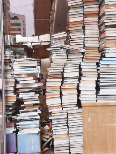 Bücher stapeln sich meterhoch auf riesigen Stapeln in einem Ladenlokal, in dem sonst nichts ist. (Gilt auch für die weiteren Bilder.)