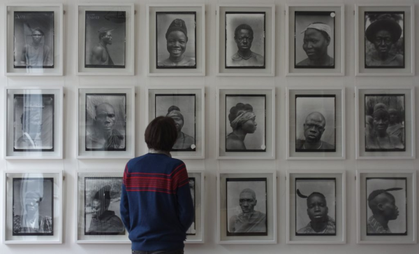 A person is looking at a wall full of enlarged photographs. The photographs are, or were, so-called 'physical type' photographs, colonial-era documentation meant to showcase general physical characteristics of (West-)African people. The photographs here, however, are presented not as showcasing a type, but as portraits of individual people. It is part of the exhibition 'Photographic affordances: revisiting N. W. Thomas’s photographic archives' at the Royal Anthropological Institute, 2018. Learn more about the organisation behind the exhibition here: https://re-entanglements.net/