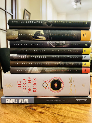 Stack of hardback books, starting with a book on weaving, then Lord of the Rings in one glorious volume, and then all the Murderbot books. 