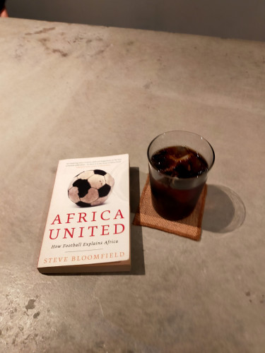 Photo is of a cement counter. On it are a white paperback book AFRICA UNITED with a soccer ball where one of the black pentagons is shaped like the continent of Africa. To the right is a glass tumbler of black iced coffee on a square brown wooden coaster